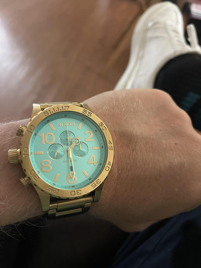 Man sitting inside and showing off his gold and turquoise Nixon 51-30 Chrono analog watch