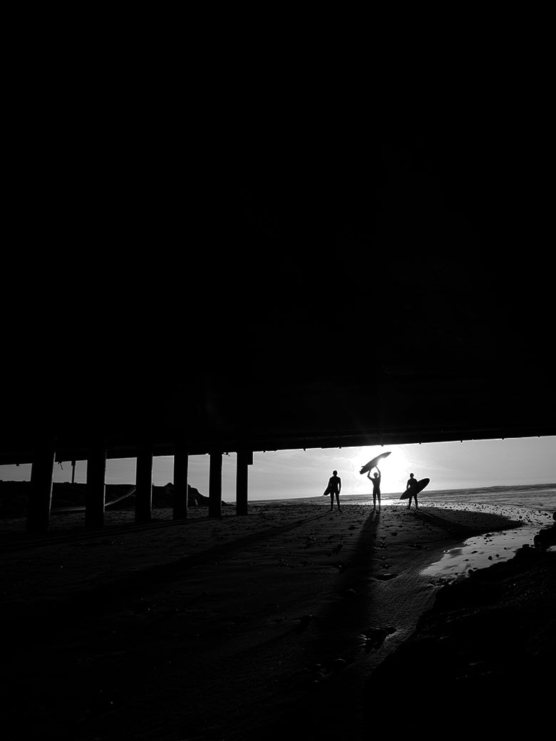 Three surfers hold their surfboards near the ocean while the sun sets